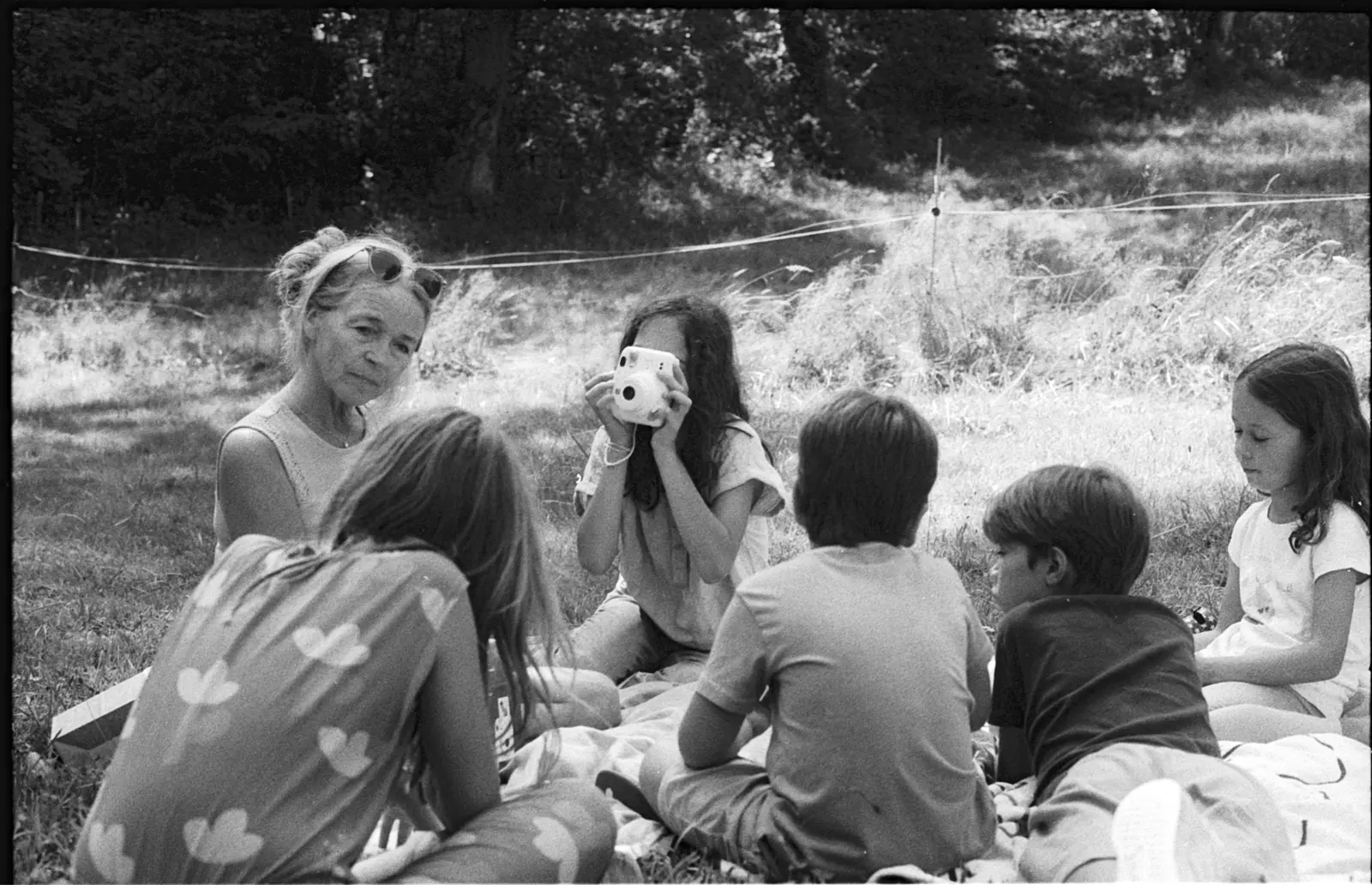 Groupe assis dans l'herbe pendant un atelier photo, jeune fille prenant une photo au Polaroid, Les Gennevriers — août 2024